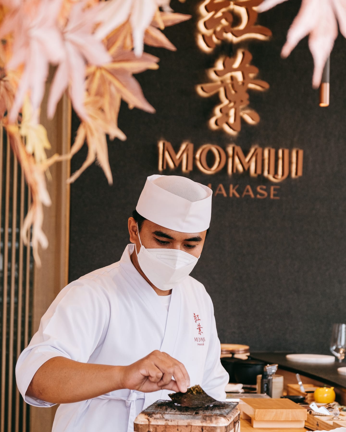 A Momiji chef preparing nori at the counter beneath the illuminated 紅葉 Momiji Omakase sign and hanging maple leaves